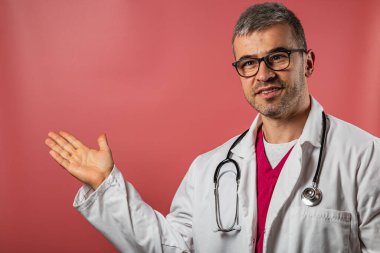 Laboratuvar önlüğü ve gözlük takan bir doktor parlak ışıklı bir odada duruyor, sağ eliyle gülümsüyor ve sanki izleyiciye önemli bir şey açıklıyormuş gibi el kol hareketi yapıyor..