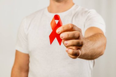Beyaz gömlekli bir adam elini uzatarak AIDS farkındalığını sembolize eden kırmızı bir kurdele sergiliyor. Ayar, mesajı vurgulayan tarafsız bir arkaplana sahip.