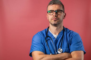 Mavi önlük ve gözlük takan bir sağlık uzmanı pembe arka planda kollarını kavuşturarak rahatça ayakta duruyor. Ciddi tavrı profesyonelliği ve kendini adamışlığı gösteriyor..