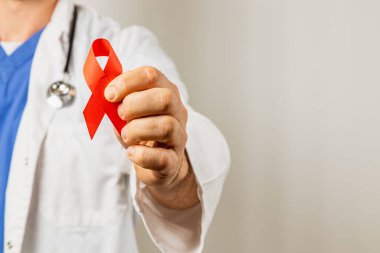 Beyaz laboratuvar önlüğü ve mavi önlük giyen bir sağlık uzmanı HIV ve AIDS 'e karşı farkındalık yaratmak için kırmızı kurdele tutuyor. Görüntü, sağlık ve desteğe olan bağlılığı vurguluyor.