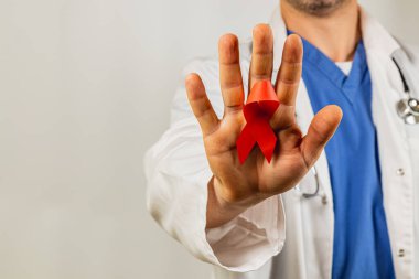 Önlüklü bir sağlık uzmanı, HIV farkındalığını sembolize eden kırmızı bir kurdeleyle el kaldırıyor. Ayar tarafsız, iletilen mesajın önemini vurguluyor.