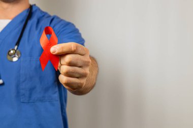 Mavi önlük giyen bir sağlık uzmanı, HIV ve AIDS farkındalığını sembolize eden kırmızı bir kurdele gösteriyor. Tarafsız arka plan dayanışmanın önemli mesajını vurguluyor.