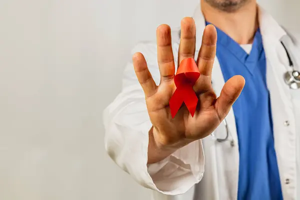 Önlüklü bir sağlık uzmanı, HIV farkındalığını sembolize eden kırmızı bir kurdeleyle el kaldırıyor. Ayar tarafsız, iletilen mesajın önemini vurguluyor.