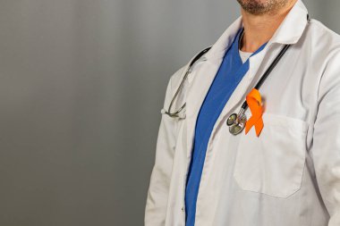 Bir sağlık çalışanı mavi önlüklerin üzerine beyaz laboratuvar önlüğü giyer, yakasında turuncu bir kurdele ile sağlık girişiminin farkındalığını arttırır..