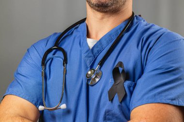 Bir sağlık çalışanı kollarını kavuşturup göğsüne siyah bir kurdele takıyor. Arka plan hafifçe bulanık, işçilerin bağlılığını vurguluyor..