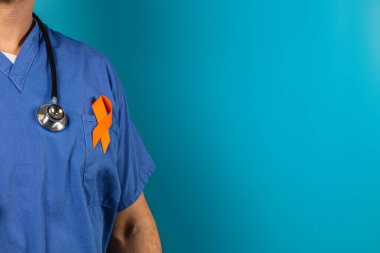 Mavi önlüklü bir tıp uzmanı, turkuaz bir arka plana karşı duruyor. Giysilerinde turuncu farkındalık şeridi sergiliyor ve sağlık nedenlerini vurguluyor..