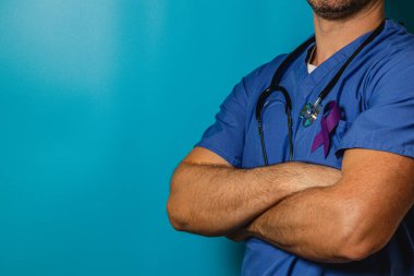 Bir sağlık uzmanı, kolları çapraz, mavi önlük ve göğsünde mor bir kurdele ile güvenle ayakta duruyor. Arkaplan, çarpıcı bir kontrastlık yaratan katı bir deniz mavisi rengidir..