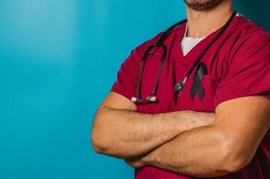 Bir sağlık çalışanı, kollarını kavuşturmuş, kestane rengi önlük ve stetoskop takmış, güven içinde ayakta duruyor. Önlüklerin üzerinde siyah bir kurdele sergileniyor, mavi bir arkaplana karşı farkındalığı ya da yasını simgeliyor..