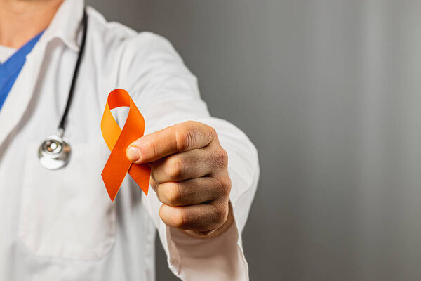 A health professional in a white coat displays an orange ribbon, symbolizing support for various health campaigns. The setting has a neutral background emphasizing the message of awareness.