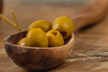 Ahşap bir kaşık, yumuşak dokuları ve canlı renklerini sergileyen olgunlaşmış yeşil zeytinleri barındırır. Köy tablosu, doğal cazibeyi arttıran sıcak bir arka plan sağlar..