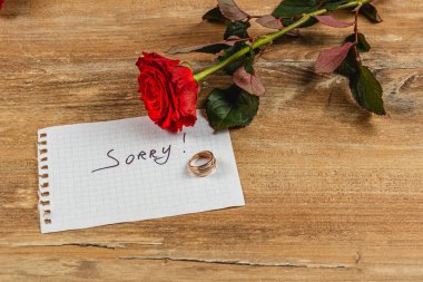 Kırmızı bir gül, ahşap bir yüzeyde altın bir yüzüğün yanında duruyor. Yanında da, üzerinde 