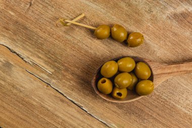 Bir ahşap kaşık yanında bir zeytin şişiyle birlikte birkaç yeşil zeytin tutar. Arka planda doğal görünümü geliştiren ahşap bir yüzey var..
