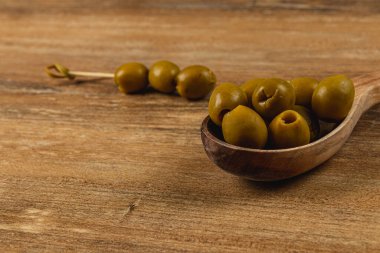 Ahşap bir kaşık, kırsal ahşap bir masa karşısında birkaç yeşil zeytin tutar. Kaşığın yanında, küçük bir demet zeytin doğal ortama eklenir, taze malzemeler sergilenir..