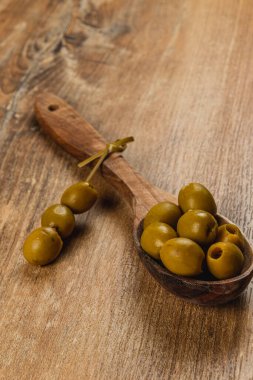 Ahşap bir kaşık, parlak dokularını gösteren birkaç yeşil zeytin tutar. Birkaç zeytin kırsal bir ahşap masaya saçılmış. Ortam doğal bir atmosfer yayıyor..