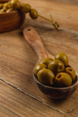 Ahşap bir kaşık kırsal bir ahşap masada birkaç yeşil zeytin tutar. Yakınlarda, daha fazla zeytin içeren bir kase taze malzemelerin ve mutfak odaklarının altını çizerek doğal ortama ekliyor..