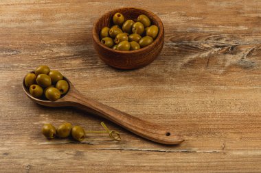 Tahta bir kaşık ve bir kâseyle birlikte yeşil zeytinler ahşap bir yüzeye dizilir. Kır manzarası malzemelerin doğal dokularını ve renklerini vurguluyor..