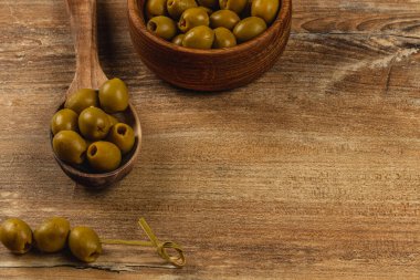 Yeşil zeytinler daha fazla zeytin ile dolu bir kasenin yanındaki ahşap bir masada düzenlenir. Ahşap bir kaşık, çeşitli zeytinler taşır, kırsal mutfak hazırlıkları için mükemmel olur..
