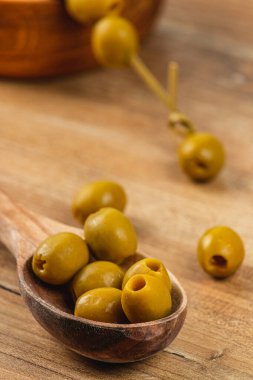 Ahşap bir kaşık kırsal bir ahşap masada küçük bir yığın yeşil zeytin tutar. Güneş ışığı zeytinlerin parlak görünümünü güçlendirir, pürüzsüz yüzeylerini ve topraksı tonlarını gösterir..