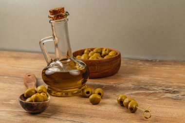 Zeytinyağıyla dolu bir cam şişe, ahşap zeytin kasesinin yanında oturur. Bazı zeytinler masaya saçılmış, kırsal mutfak temasını geliştiriyor..