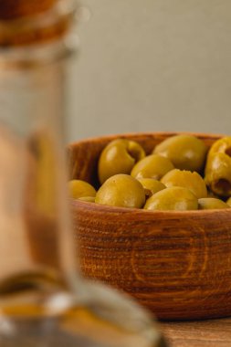 Taze yeşil zeytinlerle dolu kırsal ahşap bir kase bir masanın üzerinde otururken, bir cam şişe ön planda hafifçe bulanıklaşır ve ortamda sıcak bir ortam yaratır..