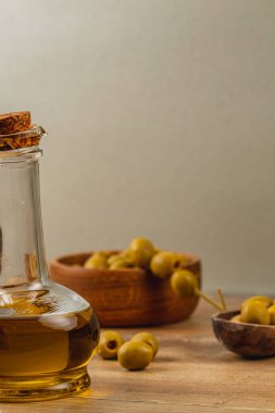 Altın zeytinyağıyla dolu bir cam şişe ahşap bir masada yeşil zeytinlerle dolu kaselerin yanında oturur ve yemek pişirmek için ideal bir kırsal mutfak ortamı yaratır..