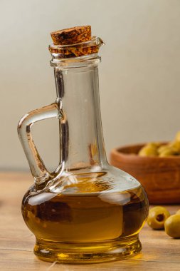 Zeytin yağıyla dolu cam bir şişe ahşap bir masanın üzerinde duruyor, mantar tıpası ile. Yakınlarda, taze yeşil zeytinler içeren bir kase, İtalyan mutfağı temasını vurguluyor..