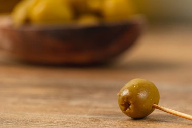Tek bir yeşil zeytin, ahşap bir masadaki kürdanın üzerinde duruyor. Arka planda, zeytin dolu bir kase biraz bulanık, kırsal bir mutfak hissi uyandırıyor..