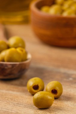 Kırsal bir ahşap yüzeyde yeşil zeytin koleksiyonu sergilenir. Bazı zeytinler küçük bir kaseye konur, davetkar ve taze bir atmosfer yaratır. Doğal ışıklandırma renkleri güçlendirir.