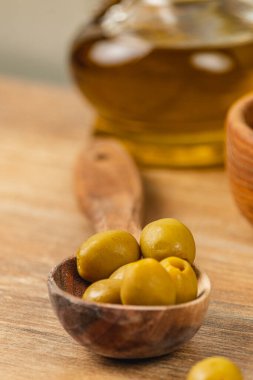 Taze yeşil zeytinler oyulmuş ahşap bir kasede dinleniyor, canlı renklerini sergiliyorlar. Arka planda bir zeytinyağı şişesi parıldıyor ve mutfak vitrinini büyütüyor..