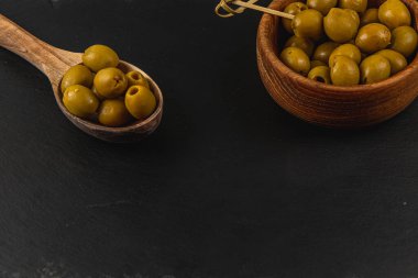 Lezzetli çukur yeşil zeytinler ahşap bir kâsede zeytin dolu bir kaşığın yanında, koyu, desenli bir yüzeye yerleştirilmiş..