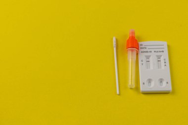 Bir COVID-19 ve grip test kiti canlı sarı bir yüzey üzerinde sergileniyor, temiz görüş için test cihazının yanına yerleştirilmiş bir çubuk ve toplama tüpü ile.