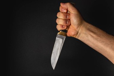 Güçlü bir el bıçağı sıkıca kavrar ve bıçağı gösterir. Karanlık arka plan sahneye yoğunlaşır, gerilim ve odaklanma hissi uyandırır..