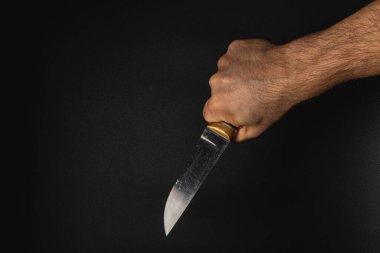 Bir el bıçağı sıkıca kavrar, bıçağın kenarını gösterir. Karanlık arka plan gerilimi vurguluyor ve el ve bıçak kombinasyonuna odaklanıyor..