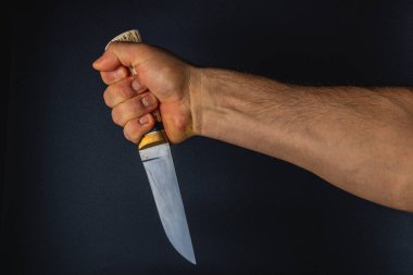 Bir el, ahşap saplı bir bıçağı sıkıca tutar, karanlık bir zemine karşı tanımlanmış kasları gösterir, dramatik bir anda hazırlık ve yoğunluğu gösterir..