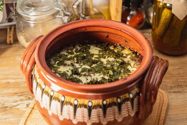 Lezzetli yeşil bir güveç ile dolu renkli bir kil tenceresi kırsal bir ahşap masada oturur. Taze otlar yemeğin kokusunu güçlendirir ve herkesi sıcaklığının tadını çıkarmaya davet eder..
