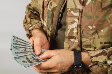Kamuflaj kıyafeti giymiş bir asker, tarafsız bir ortamda durup nakit işleme odaklı bir anı gösterirken birden fazla banknot sayar..