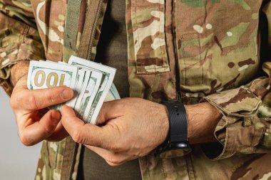 Kamuflaj üniformalı bir asker içeride dururken yüz dolarlık banknotları sayıyor. İfade, paraya odaklandığını ve niyetini gösteriyor..