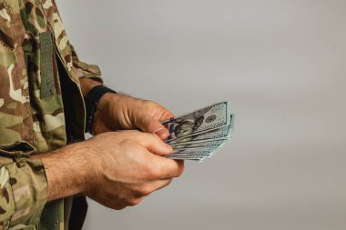 Kamuflaj üniformalı bir asker, tarafsız bir ortamda askeri yaşamla mali işlemler arasındaki zıtlığı vurgulayarak çok sayıda banknot sayıyor..