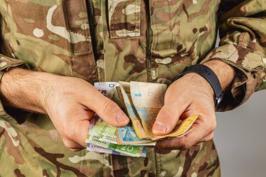 Kamuflaj üniformalı bir asker içeride çeşitli banknotları sayıyor. Odaklanmış ifade aktarım sırasında detaylara dikkat gösterir.