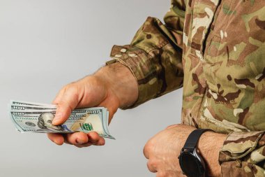 Kamuflaj teçhizatı olan bir asker birkaç yüz dolarlık banknotlar tutuyor ve para alışverişine odaklanıyor. Dekor tarafsız ve kapalı görünüyor..