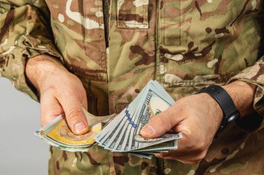 Bir asker, askeri kıyafetler giyerken elindeki parayı kontrol eder ve gündelik ışıklandırma koşulları altında çeşitli banknotların karışımını gösterir..