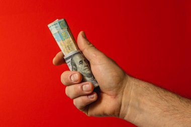 Bir el sıkıca yuvarlanmış banknotları kavrıyor, parlak kırmızı bir zemin üzerinde belirgin bir konum, kontrastı vurgulayarak ve paraya odaklanıyor.