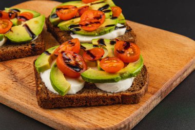 Krem peynirle kaplı koyu ekmek dilimleri, taze avokado ve iştah açıcı bir şekilde hazırlanmış canlı vişneli domatesler. Hafif bir öğle yemeği veya atıştırmalık için ideal..