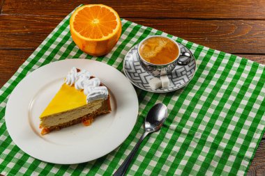 Sarı jöleli ve kremalı kremalı kremalı bir dilim kremalı kek bir fincan kahve ve canlı bir portakalın yanında beyaz bir tabağa yerleştirilir. Damalı yeşil ve beyaz masa örtüsü cazibe katıyor..