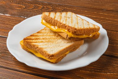 İki mükemmel ızgara peynirli sandviç, ahşap bir masanın üzerinde beyaz bir tabakta oturuyorlar. Kahvaltı ya da brunch için ideal, bu lezzetli atıştırmalıklarda altın tost ekmeği ve erimiş peynir var..