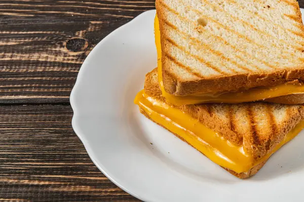 Kusursuz bir ızgara peynirli sandviç beyaz bir tabakta servis edilir, erimiş peyniri iki dilim altın rengi tost ekmeğinin arasında sergiler. Öğle yemeği, brunch ya da lezzetli bir atıştırmalık için ideal..