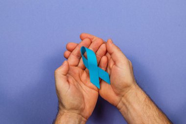 Eller, farkındalığı ve kanser tespit girişimlerine verilen desteği sembolize eden sağlam bir arka plana karşı mavi kurdele tutuyor. Bu görsel, kanser bakımı ve önlenmesinin önemini vurgular.