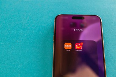03.26.25 Lviv Ukrayna Akıllı telefon ekranı mağaza uygulamalarını gösteriyor. Temu ve AliExpress için simgeler bir uyarı rozetinin yanında görünür. Cihaz bir deniz mavisi yüzeyinde duruyor..