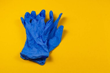 Bir çift mavi tek kullanımlık eldiven parlak sarı bir yüzeye dayanır, basit ve çarpıcı bir kompozisyonda hijyen ve temizlik temalarını önerir..