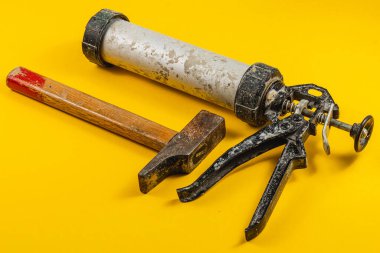 Bir çekiç, kabartma tabancası ve bir tüp dolgu malzemesi, çeşitli inşaat işleri için gerekli araçları gösteren canlı sarı bir arka planda sergilenmektedir..